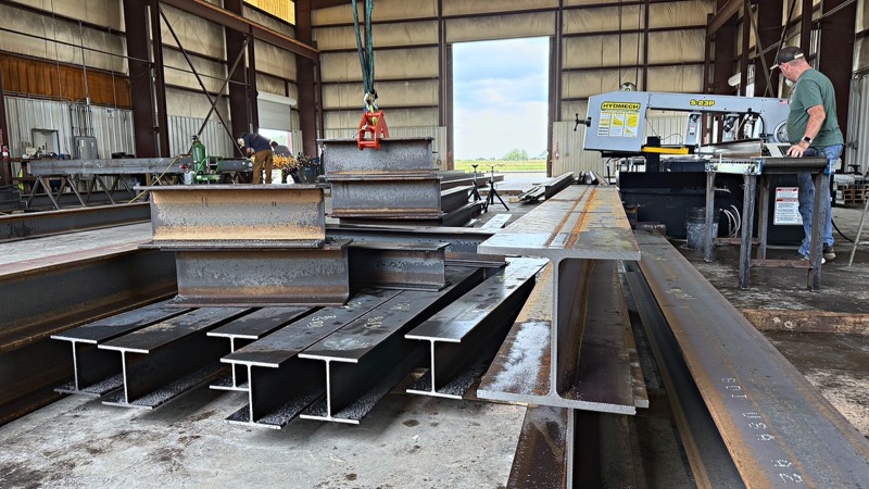Wide-flange beams stacked on the shop floor next to a band saw